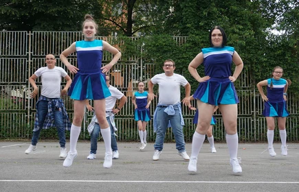 Kilkoro dziewcząt i chłopców w niebieskich strojach tanecznych w stylu czirliderek tańczy na szkolnym boisku sportowym.